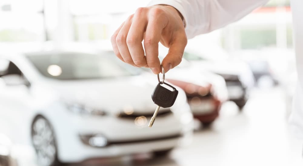Hand holding car keys in a dealership setting with blurred vehicles in the background.
