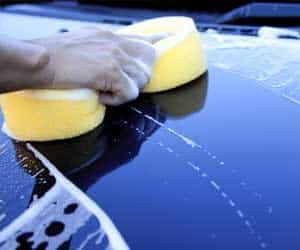 Hand washing a car with a yellow sponge, creating soapy suds on the car's surface.