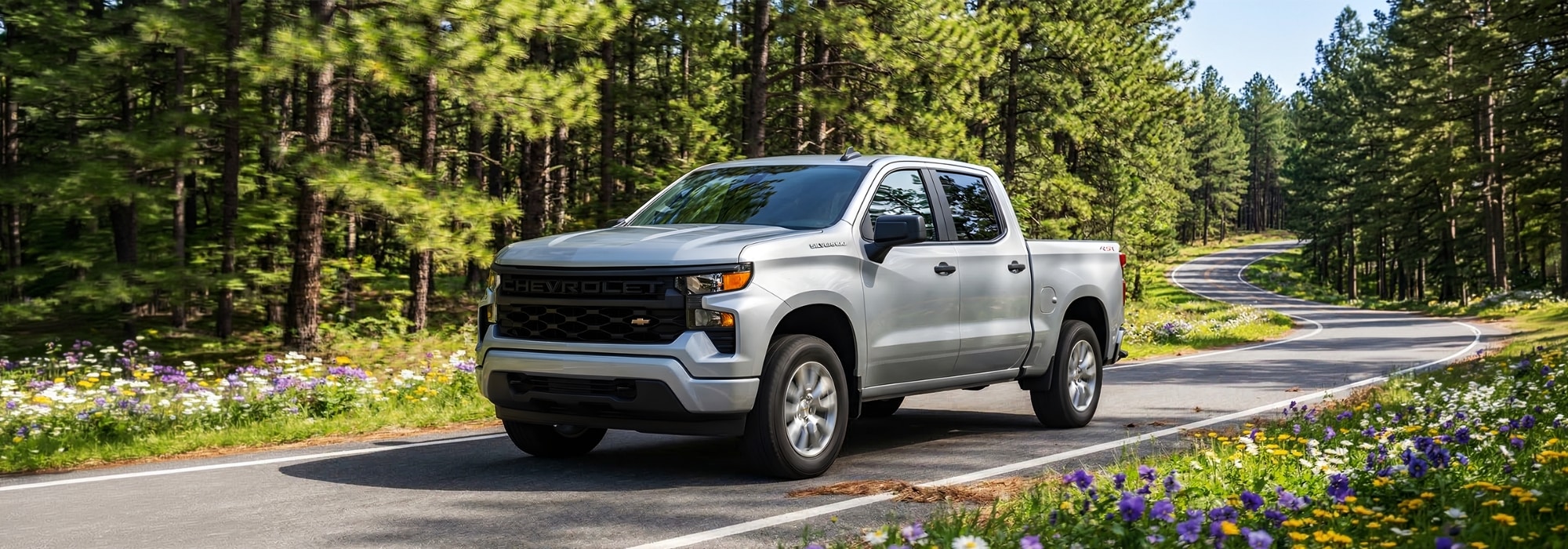 Silver Chevrolet Silverado driving on a scenic forest road lined with wildflowers.
